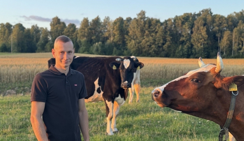 Attēlā redzams vīrietis tumšā tkreklā un divas govis. Aplārt koki un zaļa pļava.