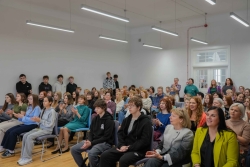 Attēlā redzami cilvēki, kas sēž zālē, dzied un applaudē