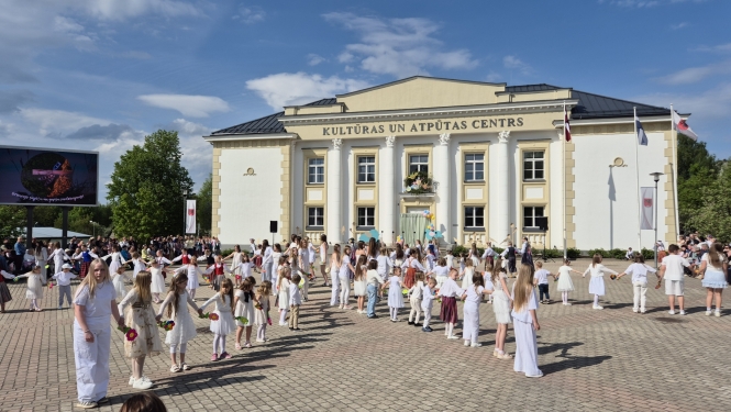 Attēlā redzams Balvu Kultūras un atpūtas centrs. Priekšplānā uz bruģa stāv sastājušies daudz bērni, ģērbušies baltā apģērbā.