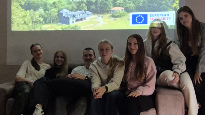 Jauniešu grupa sēž uz dīvāna telpā pie projektora ekrāna ar prezentāciju par brīvprātīgo darbu Gruzijā.