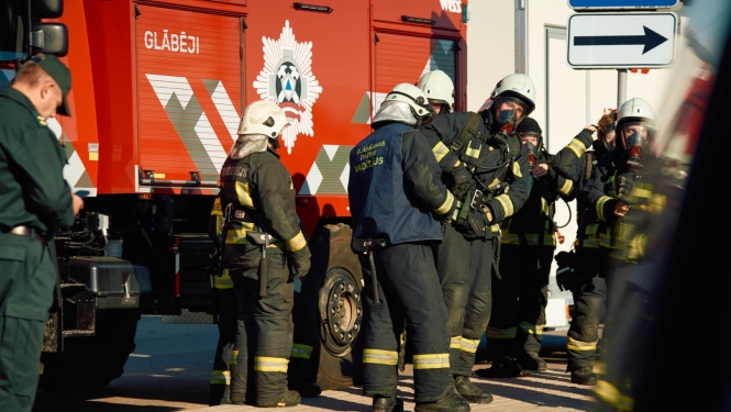 Ugunsdzēsēji glābēji aizsargtērpos pulcējušies pie sarkanas VUGD automašīnas mācību laikā. Attēlā redzami vairāki glābšanas dienesta darbinieki ekipējumā, gatavojoties uzdevumu veikšanai pilsētvidē.