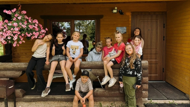 Attēlā redzami bērni, kuri pozē fotogrāfam. Daži uzsēdušies uz koka margām, citi stāv kājās. Kreisajā pusē redzams rozā petūniju pods. Fonā koka ēka.