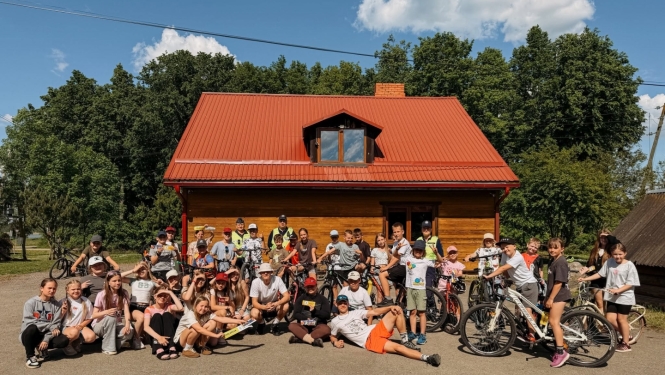 Attēlā redzami ļoti daudzi bērni un jaunieši ar velosipēdiem, pie Viļakas jauniešu mājas. Vasarīga, silta noskaņa, zilas debesis un zaļi koki fonā.