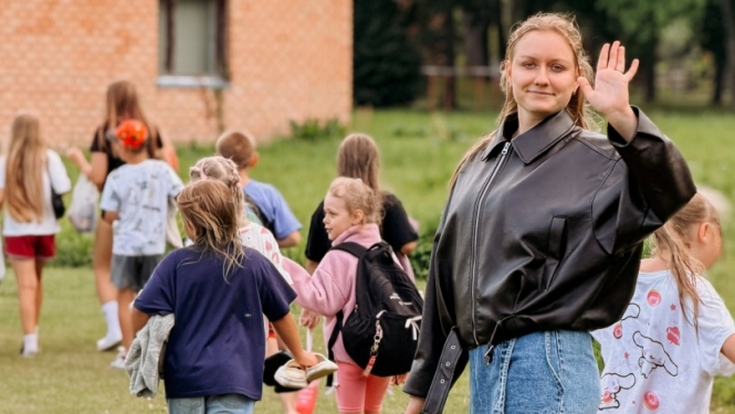 Priekšplānā redzama meitene, kas māj. Tālāk redzams bērnu bariņš no mugurpuses ar somām plecos. Fonā sarkanu ķieģeļu ēka.