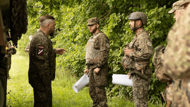 Militārpersonas dienesta formas tērpos ar Latvijas karoga un vienību atpazīšanas zīmēm atrodas pie zaļa meža mežmalas. Viens no karavīriem žestikulējot runā ar diviem kolēģiem pilnā ekipējumā (bruņuvestē un ķiverē), kuri rokās tur sarullētas kartes vai dokumentus.