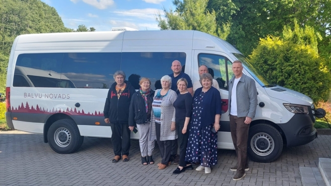 Cilvēku grupa pozē kopbildei pie balta mikroautobusa, uz kura sāniem ir uzraksts "BALVU NOVADS" un sarkans meža silueta krāsojums. Transportlīdzeklis ir pieslēgts uzlādes stacijai pie ķieģeļu ēkas.