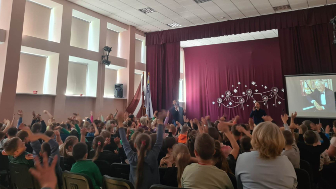 Attēlā redzama aktu zāle ar daudziem skolēniem. Skatuvi rotā bordo krāsas aizkari, un tās priekšā stāv divi vīrieši. Labajā pusē redzams ekrāns uz kura tiek projecēta Raimonda Paula bilde.