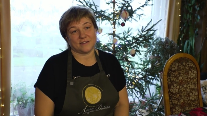 Sieviete tumšā priekšautā ar logotipu "Arnitas Labsajūtu Darbnīca" sēž telpā, kur fonā redzama izgreznota Ziemassvētku eglīte un logs.