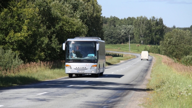 : Sudrabots Setra markas pasažieru autobuss brauc pa asfaltētu lauku ceļu saulainā dienā. Tālumā priekšā redzams balts mikroautobuss, un ceļa abās malās ir zaļi koki un pļavas.