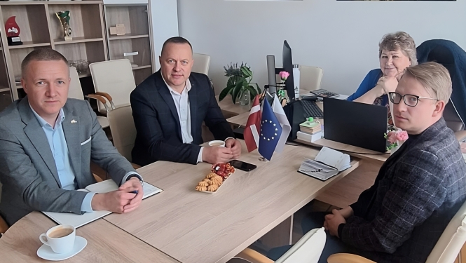 Attēlā redzami četri cilvēki, kas sēž pie galda un slatās kamerā