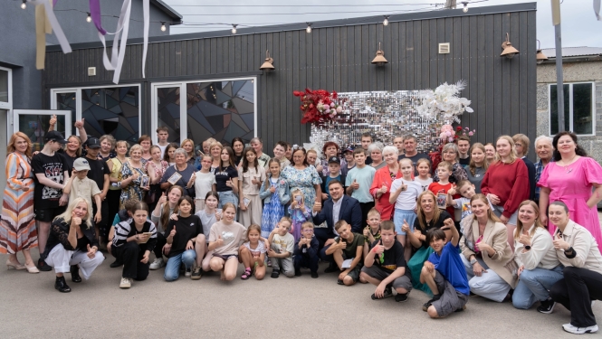 Attēlā redzama liela cilvēku grupa — bērni, jaunieši un pieaugušie — kas sapulcējušies pie ēkas kopīgai fotogrāfijai. Daudzi smaida un rāda paceltu īkšķi. Fonā redzama dekorēta siena ar ziediem un spīdīgiem elementiem, virs galvām izkārtotas lampiņas un lentes, radot svinīgu un draudzīgu pasākuma atmosfēru.