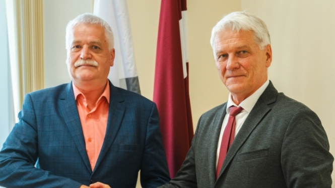 Domes priekšsēdētājs Jānis Trupovnieks spiež roku svinīgā gaisotnē telpā pie karogiem domes deputātam Imantam Slišānam.