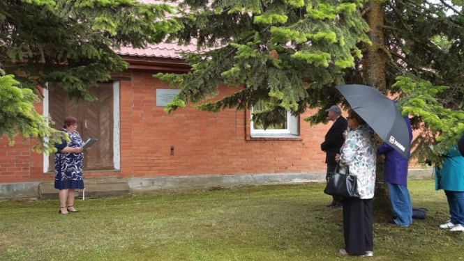 Komunistiskā genocīda upuru piemiņas brīdis pie sarkano ķieģeļu ēkas, kur pie sienas piestiprināta piemiņas plāksne. Kreisajā pusē sieviete nolasa uzrunu, bet labajā pusē koku ēnā stāv piemiņas pasākuma dalībnieki, daži no tiem patvērušies zem lietussarga.