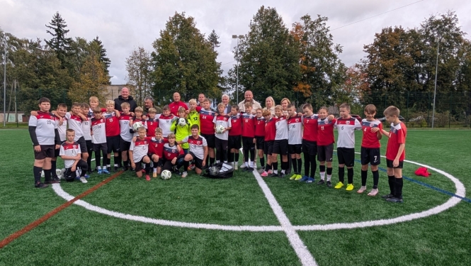 Liela bērnu futbola komanda un pieaugušie oficiālā apģērbā un sporta tērpos stāv uz jaunā futbola laukuma mākslīgā seguma stadiona atklāšanas pasākumā.