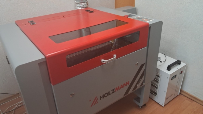Attēlā redzama lāzergravēšanas un lāzergriešanas iekārta ar zīmolu HOLZMANN. Tai ir aizsargkorpuss ar stikla skatlodziņu, ventilācijas izvads dūmu novadīšanai un blakus novietota dzesēšanas iekārta, kas nodrošina lāzera darbībai nepieciešamo dzesēšanu.