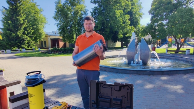 Arnis stāv parkā pie strūklakas un tur rokās lielu, cilindrisku metāla urbi. Priekšplānā uz koka soliņa izvietots profesionāls urbšanas aprīkojums dzeltenā un melnā krāsā.