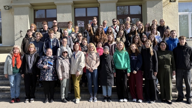 Attēlā redzama liela cilvēku grupa, kas pozē kopīgai fotogrāfijai pie ēkas ieejas. Grupā ir dažāda vecuma cilvēki – gan jaunieši, gan pieaugušie. Visi stāv uz kāpnēm un laukuma ēkas priekšā saulainā dienā. Daudzi ir ģērbušies jakās un mēteļos, kas liecina par vēsāku laiku. Fotogrāfija izskatās kā skolas, organizācijas vai kopienas pasākuma kopbilde.