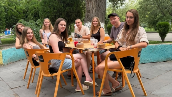 Attēlā redzami vairāki jaunieši, sēžot pie galda. Uz zemes guļ gaišs liels suns.