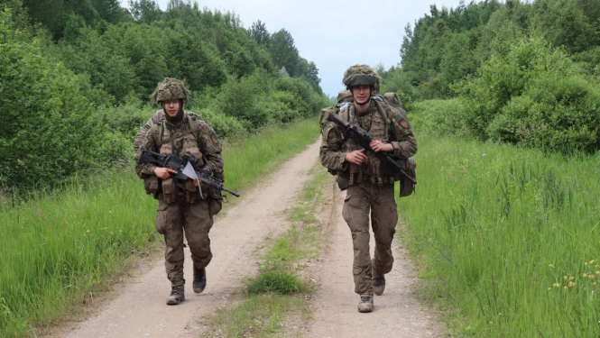 Divi karavīri pilnā kaujas ekipējumā un ar triecienšautenēm rokās iet pa meža smilts ceļu apmācību laikā.
