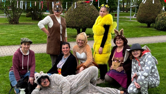 Grupa cilvēku parkā pozē kopīgai fotogrāfijai, tērpušies dažādos dzīvnieku un pasaku tēlu kostīmos. Daļa sēž un tup zālienā, citi stāv, radot rotaļīgu un priecīgu noskaņu. Fonā redzami dekorējumi un sakopts parks. Blakus grupai atrodas arī suns.
