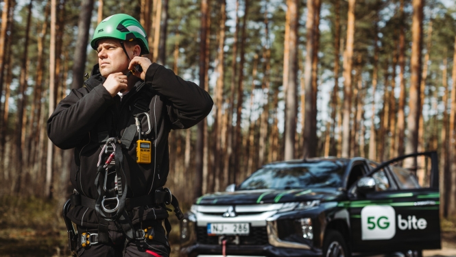 "Bite" telekomunikāciju inženieris darba ekipējumā un zaļā aizsargķiverē gatavojas darbam meža apvidū. Fonā redzams apvidus auto ar "5G bite" zīmolu.
