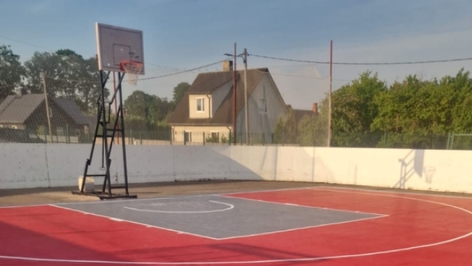 Āra basketbola laukums ar spilgti sarkanu un pelēku moduļu segumu. Laukumā redzams basketbola grozs, un to ieskauj balta sēta.