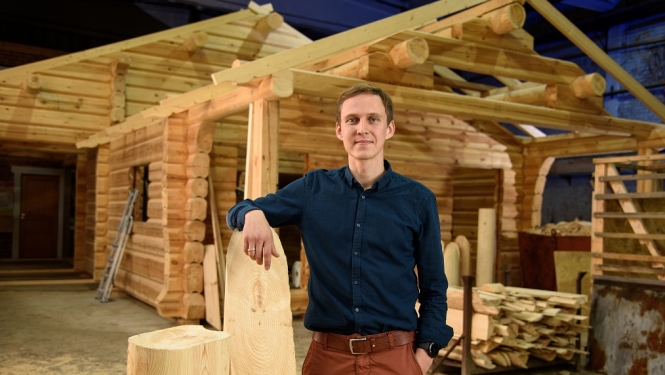 Jānis Igaunis tumši zilā kreklā pozē ražošanas angārā uz nepabeigtas guļbūves koka mājas karkasa fona, atbalstījis roku pret lielu koka brusu.
