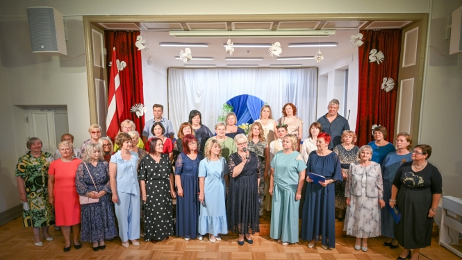 Attēlā redzama liela cilvēku grupa, kas stāv uz skatuves svinīgā pasākumā. Priekšplānā vairākas sievietes ir saposušās svētku tērpos, viena no viņām runā mikrofonā, bet cita tur zilu mapi. Fonā redzami sarkani aizkari, balti dekorējumi un Latvijas karogs, kas rada oficiālu un svinīgu noskaņu. Visi ir pavērsti pret skatītājiem, iespējams, kopīgai uzstāšanās reizei vai piemiņas foto.