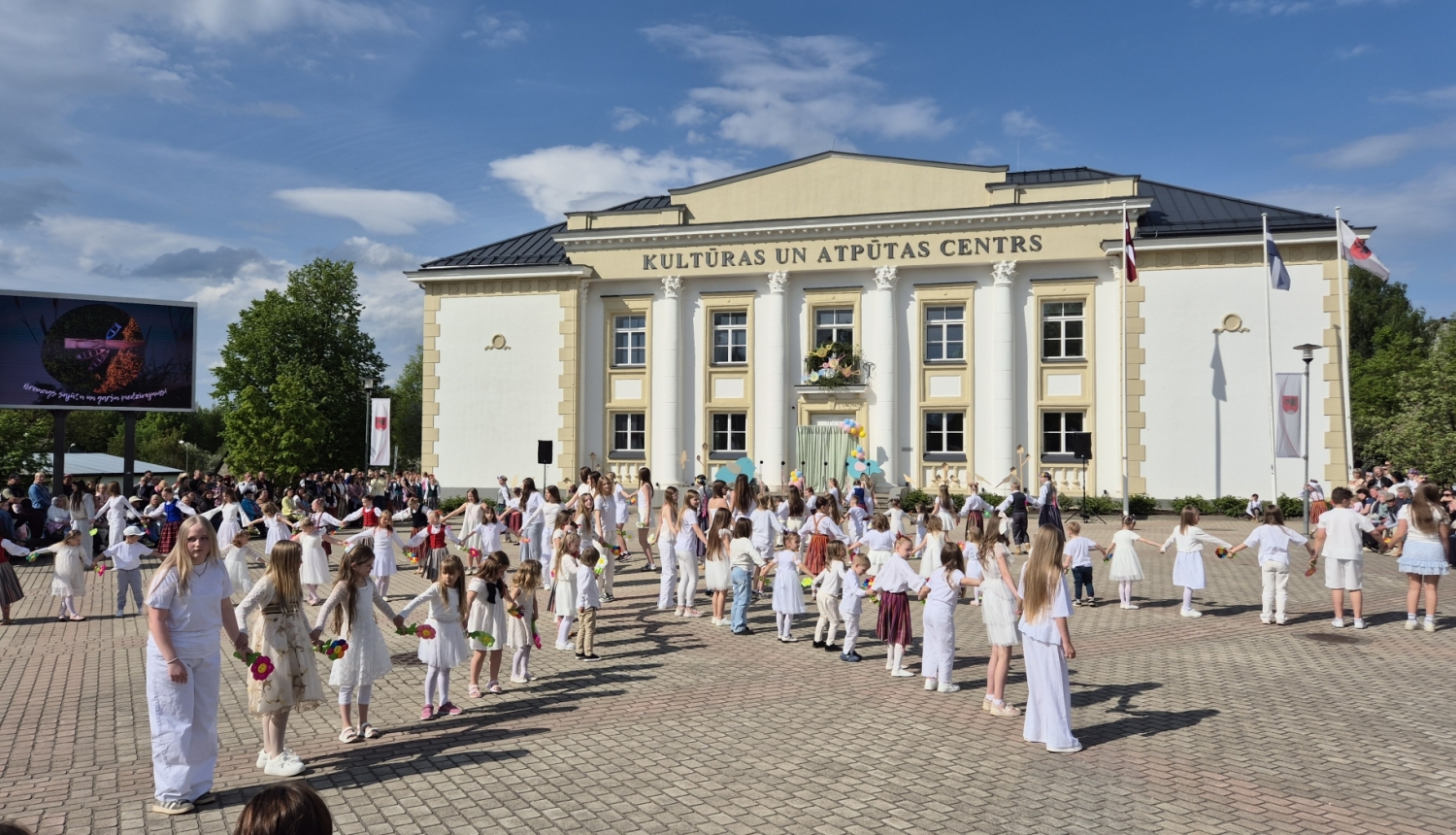 Attēlā redzams Balvu Kultūras un atpūtas centrs. Priekšplānā uz bruģa stāv sastājušies daudz bērni, ģērbušies baltā apģērbā.