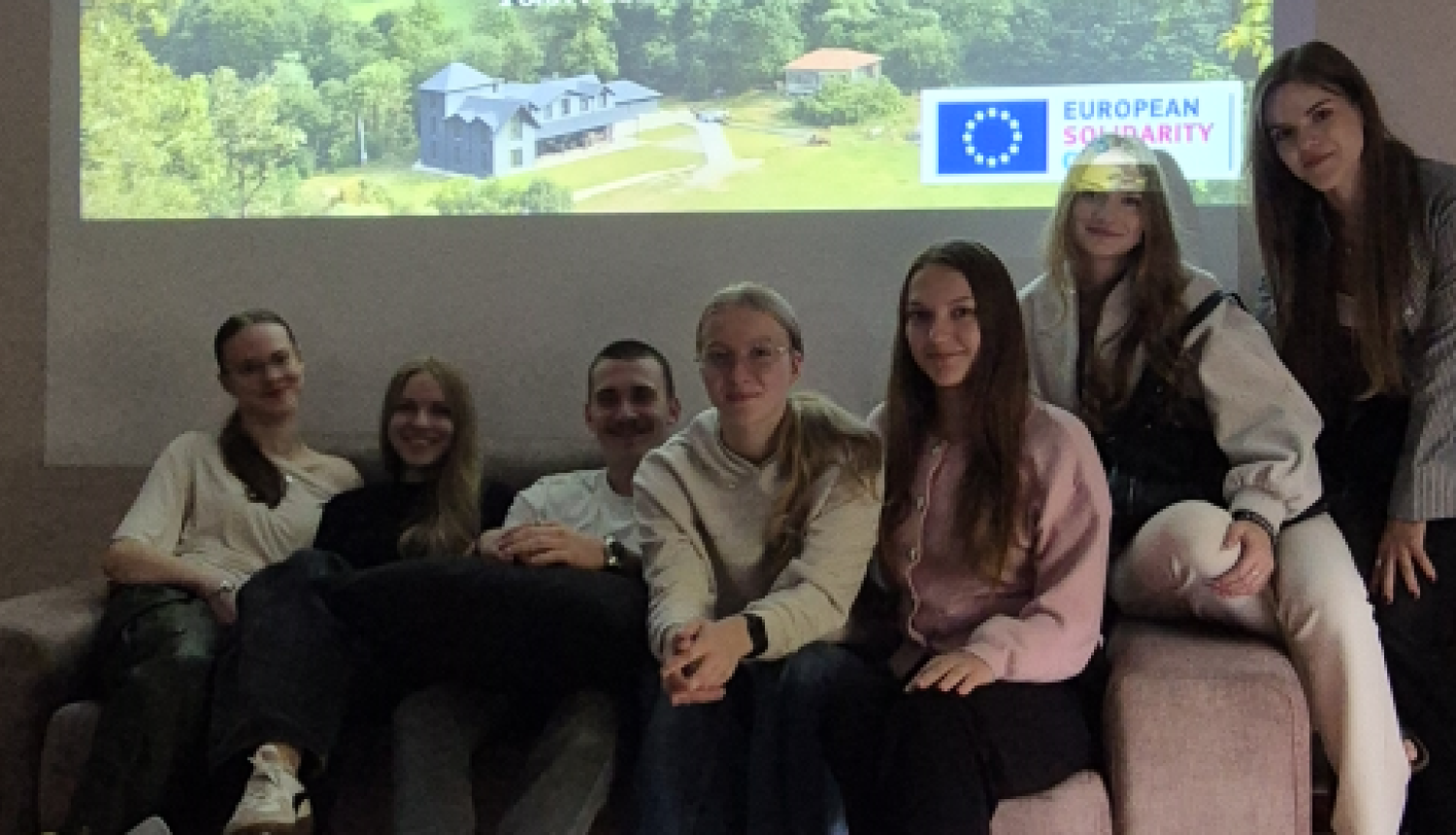 Jauniešu grupa sēž uz dīvāna telpā pie projektora ekrāna ar prezentāciju par brīvprātīgo darbu Gruzijā.