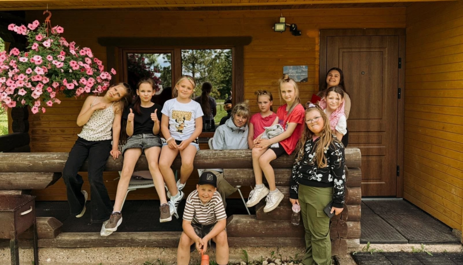 Attēlā redzami bērni, kuri pozē fotogrāfam. Daži uzsēdušies uz koka margām, citi stāv kājās. Kreisajā pusē redzams rozā petūniju pods. Fonā koka ēka.