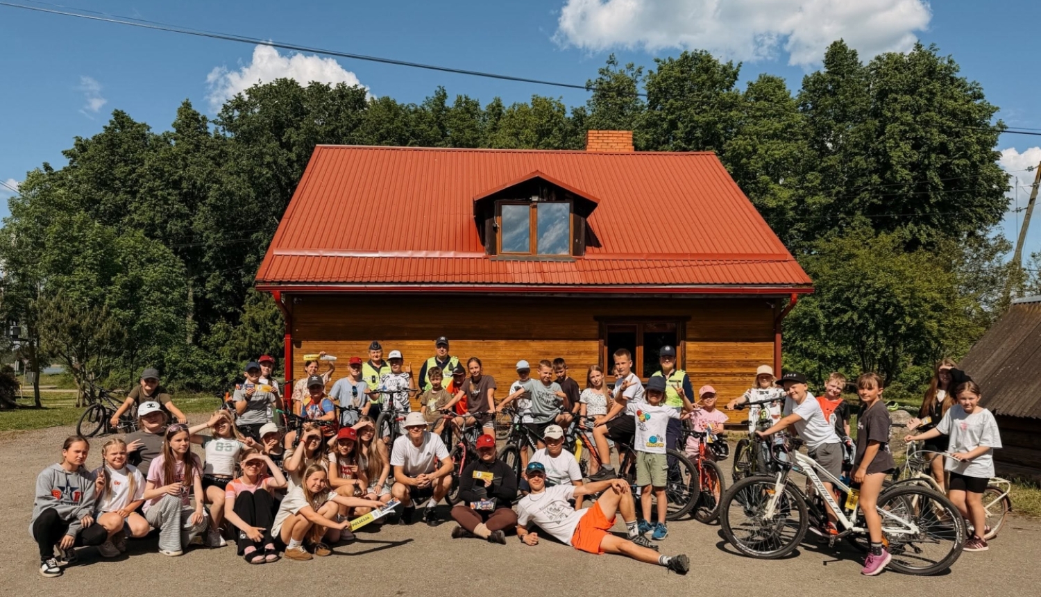 Attēlā redzami ļoti daudzi bērni un jaunieši ar velosipēdiem, pie Viļakas jauniešu mājas. Vasarīga, silta noskaņa, zilas debesis un zaļi koki fonā.