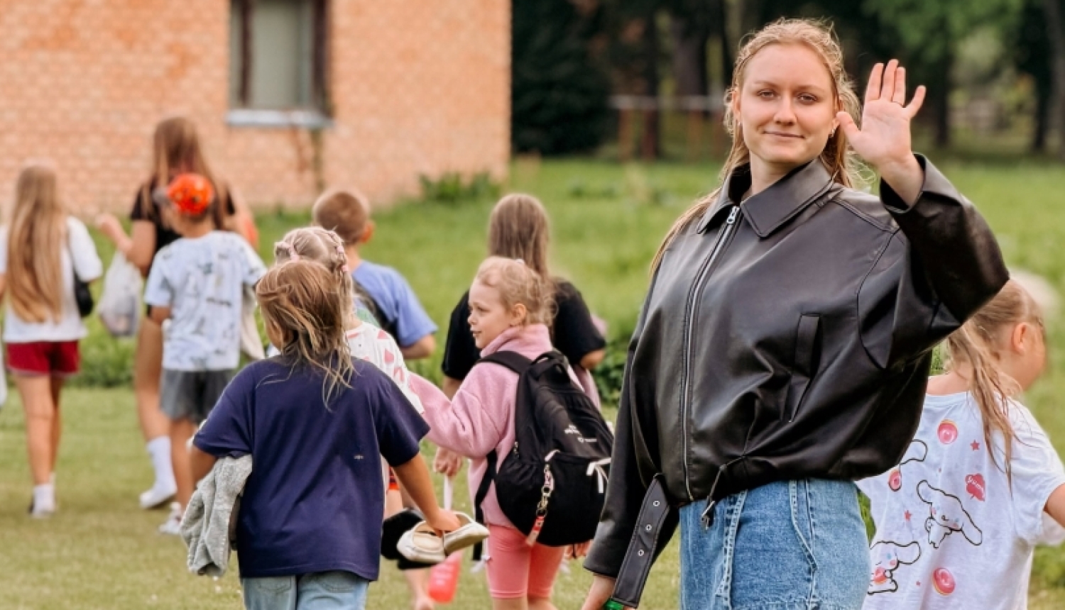 Priekšplānā redzama meitene, kas māj. Tālāk redzams bērnu bariņš no mugurpuses ar somām plecos. Fonā sarkanu ķieģeļu ēka.