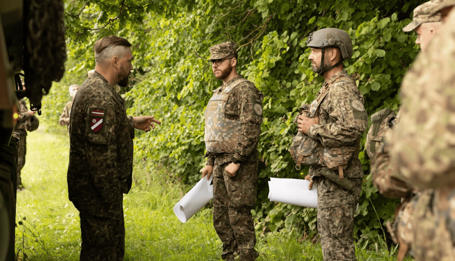 Militārpersonas dienesta formas tērpos ar Latvijas karoga un vienību atpazīšanas zīmēm atrodas pie zaļa meža mežmalas. Viens no karavīriem žestikulējot runā ar diviem kolēģiem pilnā ekipējumā (bruņuvestē un ķiverē), kuri rokās tur sarullētas kartes vai dokumentus.