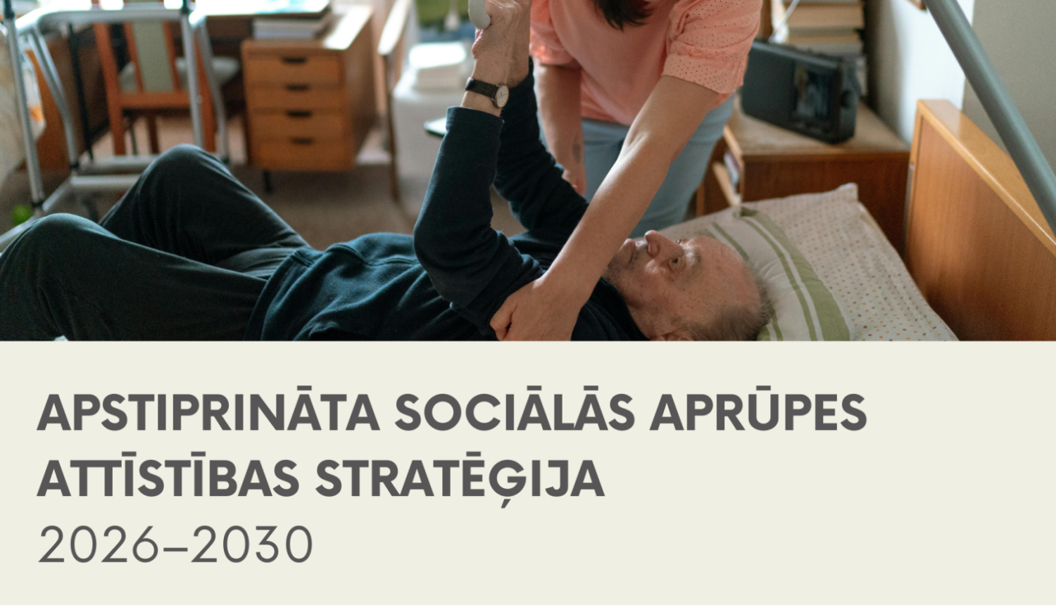 Informatīvs attēls ar uzrakstu "APSTIPRINĀTA SOCIĀLĀS APRŪPES ATTĪSTĪBAS STRATĒĢIJA 2026–2030". Augšpusē redzama fotogrāfija, kurā sociālā darbiniece jeb aprūpētāja palīdz gulošam vecāka gadagājuma vīrietim gultā, turot viņu aiz rokas un palīdzot piecelties.