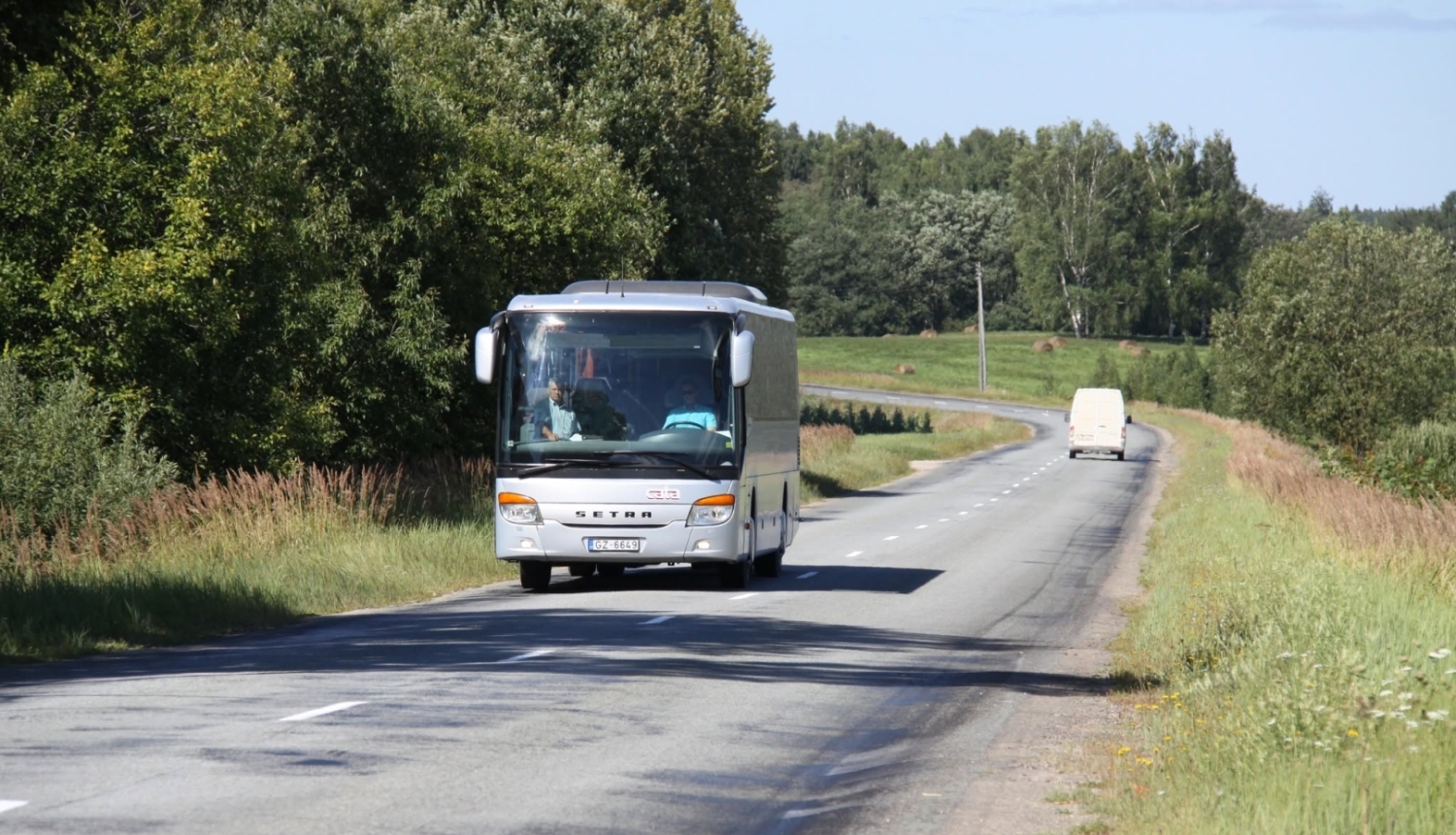 : Sudrabots Setra markas pasažieru autobuss brauc pa asfaltētu lauku ceļu saulainā dienā. Tālumā priekšā redzams balts mikroautobuss, un ceļa abās malās ir zaļi koki un pļavas.