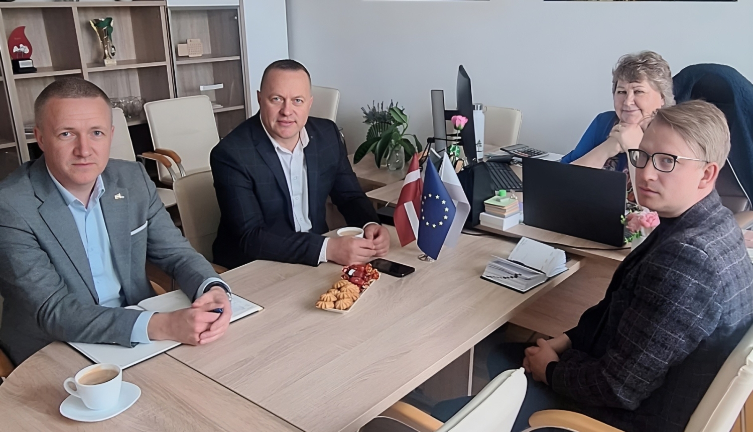 Attēlā redzami četri cilvēki, kas sēž pie galda un slatās kamerā