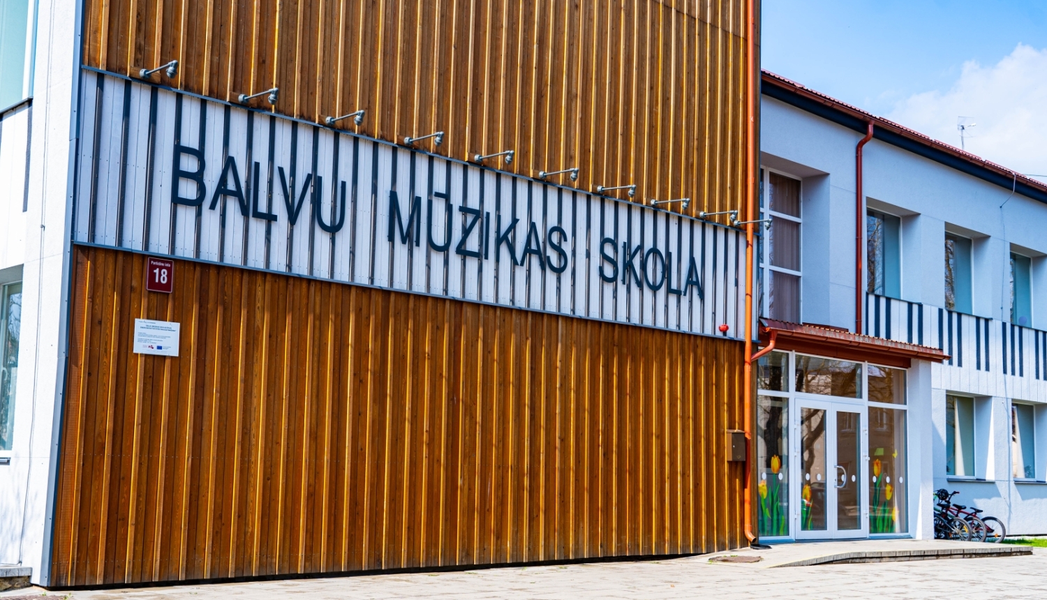 Attēlā redzama Balvu Mūzikas skolas fasāde. Gaiša ēka ar koka elementiem un skolas uzrakstu.