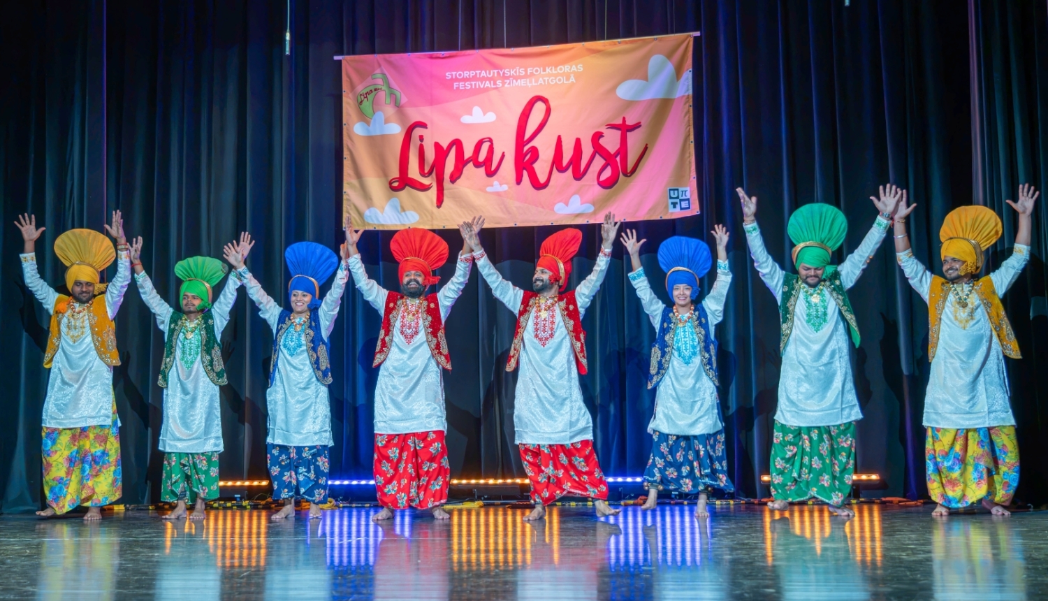 Uz skatuves rindā stāv astoņi tautastērpos ģērbušies dejotāji ar paceltām rokām. Viņi valkā košus, krāsainus tērpus un lielas tradicionālas galvassegas, radot svinīgu un enerģisku noskaņu. Fonā redzams festivāla plakāts un tumši skatuves aizkari, bet grīdu izgaismo skatuves apgaismojums.