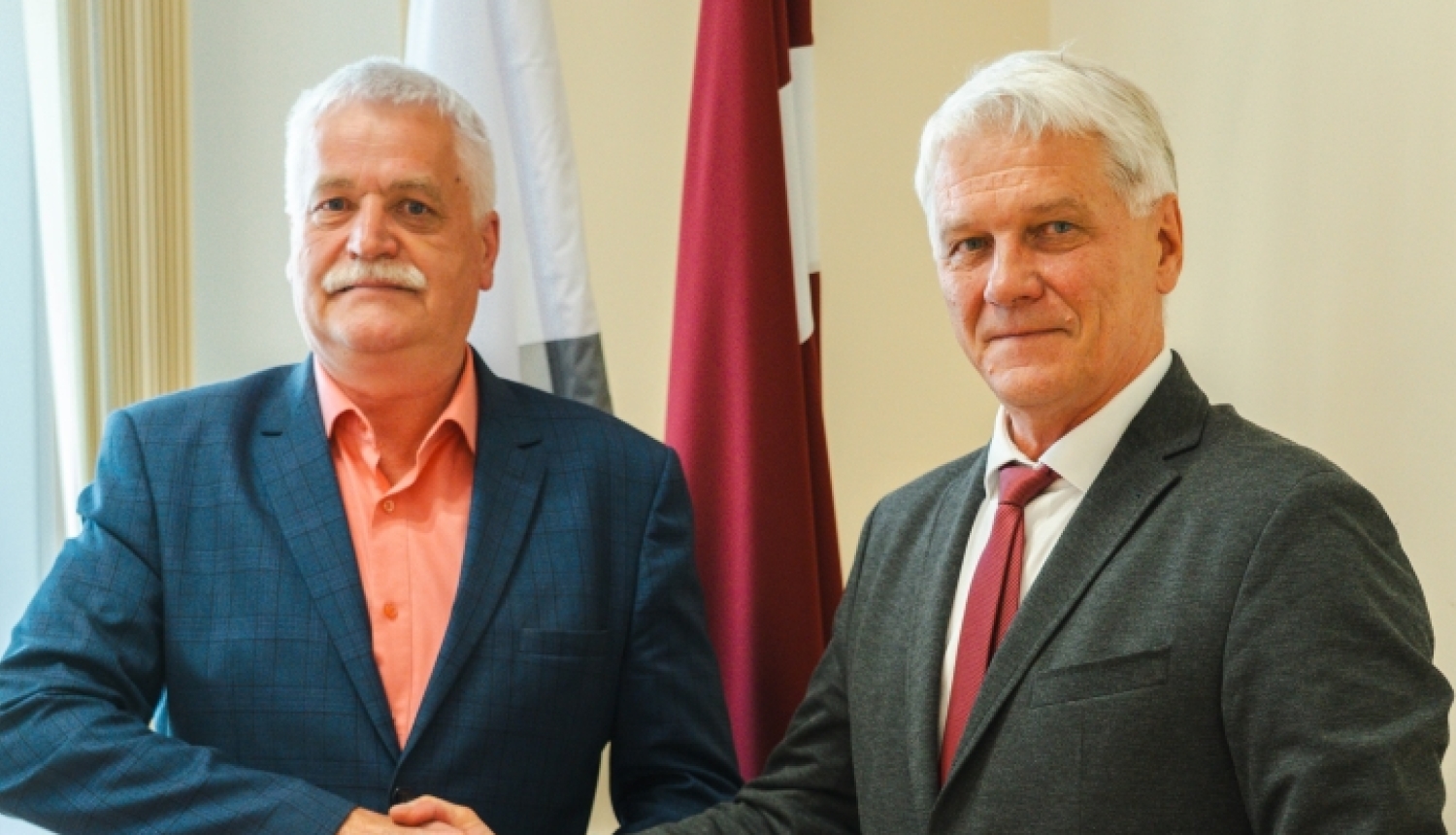 Domes priekšsēdētājs Jānis Trupovnieks spiež roku svinīgā gaisotnē telpā pie karogiem domes deputātam Imantam Slišānam.