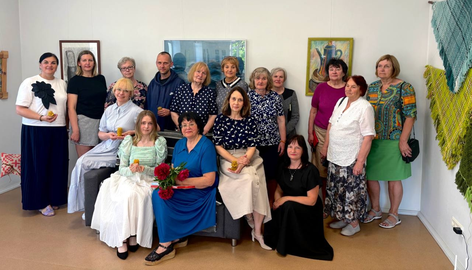 Cilvēku grupa pozē kopbildei izstādes atklāšanas pasākumā bibliotēkā. Fonā pie baltajām sienām redzamas gleznas un labajā pusē – austi tekstildarbi. Priekšplānā sēdošajai sievietei zilā kleitā rokās ir sarkanas peonijas.