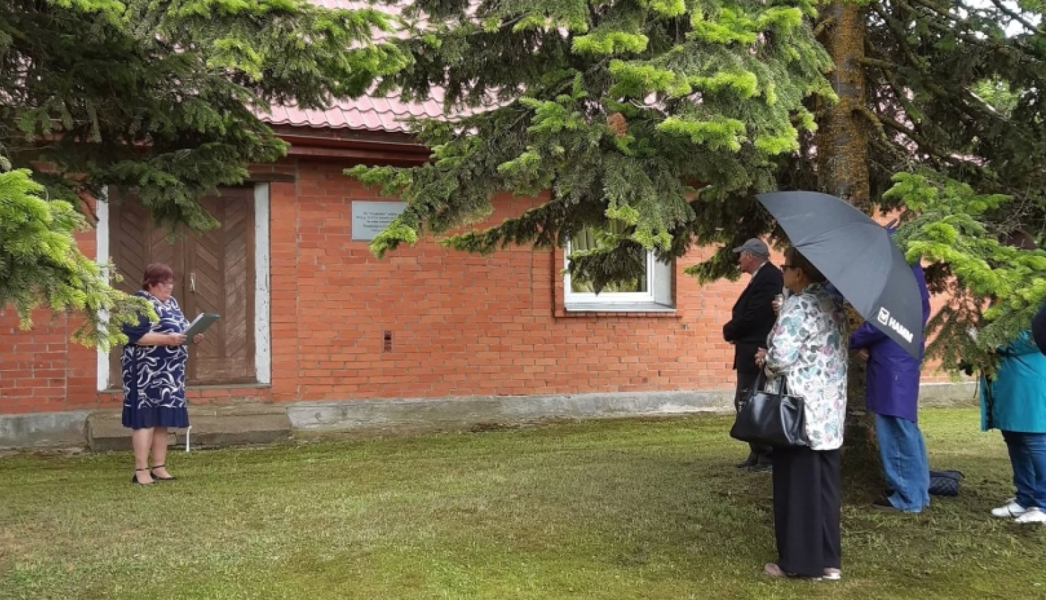 Komunistiskā genocīda upuru piemiņas brīdis pie sarkano ķieģeļu ēkas, kur pie sienas piestiprināta piemiņas plāksne. Kreisajā pusē sieviete nolasa uzrunu, bet labajā pusē koku ēnā stāv piemiņas pasākuma dalībnieki, daži no tiem patvērušies zem lietussarga.