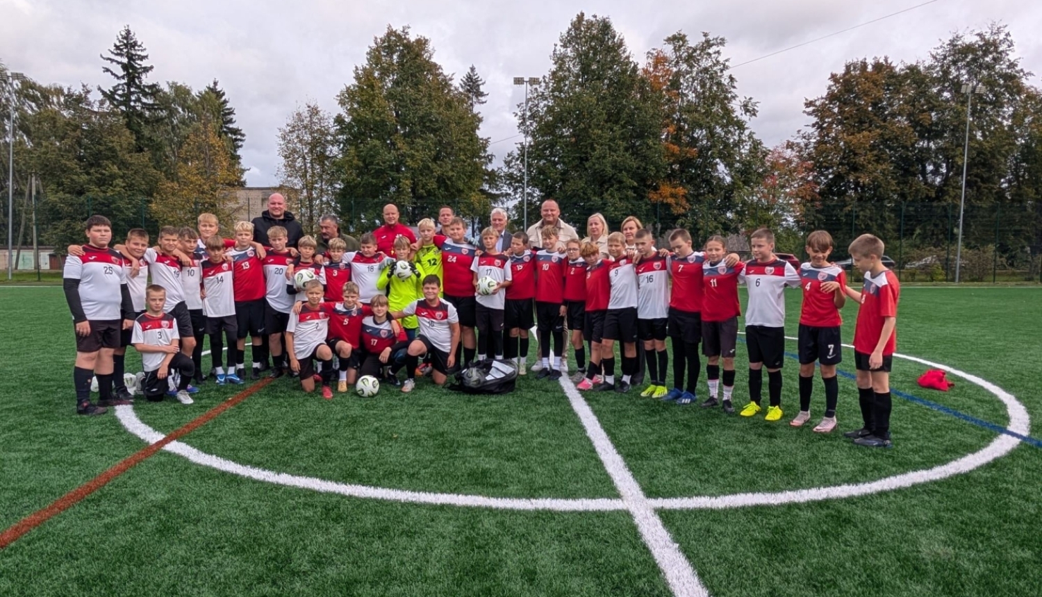 Liela bērnu futbola komanda un pieaugušie oficiālā apģērbā un sporta tērpos stāv uz jaunā futbola laukuma mākslīgā seguma stadiona atklāšanas pasākumā.