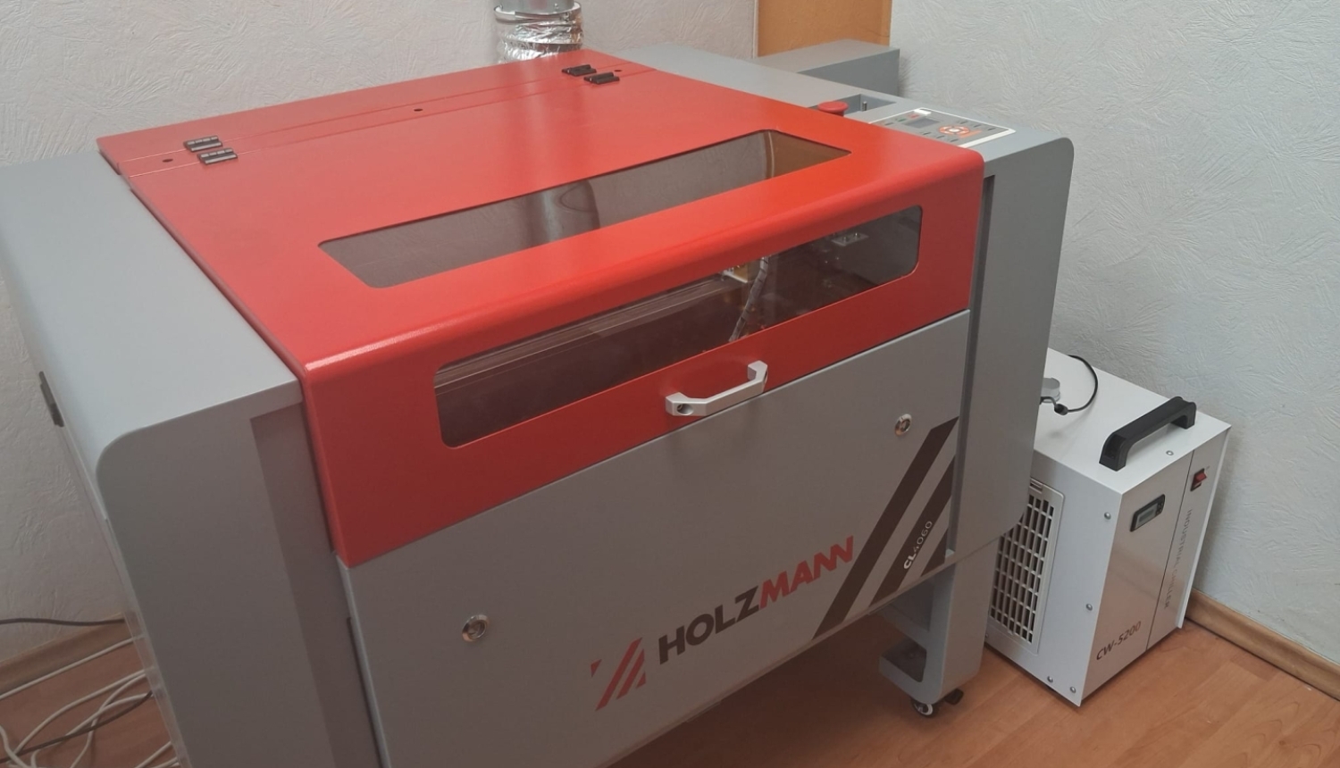 Attēlā redzama lāzergravēšanas un lāzergriešanas iekārta ar zīmolu HOLZMANN. Tai ir aizsargkorpuss ar stikla skatlodziņu, ventilācijas izvads dūmu novadīšanai un blakus novietota dzesēšanas iekārta, kas nodrošina lāzera darbībai nepieciešamo dzesēšanu.