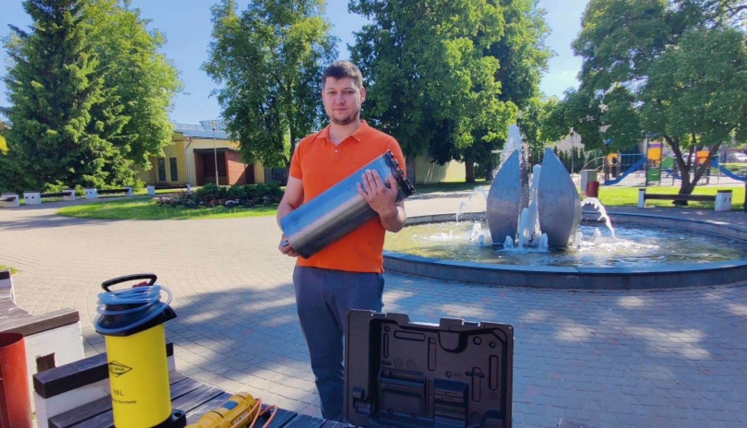 Arnis stāv parkā pie strūklakas un tur rokās lielu, cilindrisku metāla urbi. Priekšplānā uz koka soliņa izvietots profesionāls urbšanas aprīkojums dzeltenā un melnā krāsā.