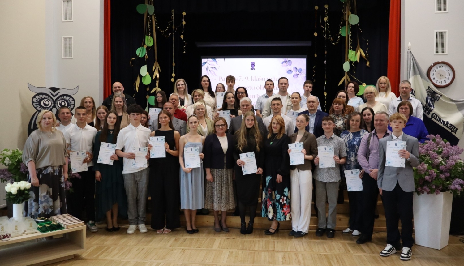 Attēlā redzama liela cilvēku grupa skolas vai ģimnāzijas aktu zālē. Daudzi jaunieši tur rokās diplomus vai apliecības, kas liecina par izlaidumu vai apbalvošanas pasākumu. Fonā ir skatuve ar dekorācijām, bet priekšplānā redzami ziedi un pedagogi, vecāki vai viesi, kas piedalās svinīgajā notikumā.