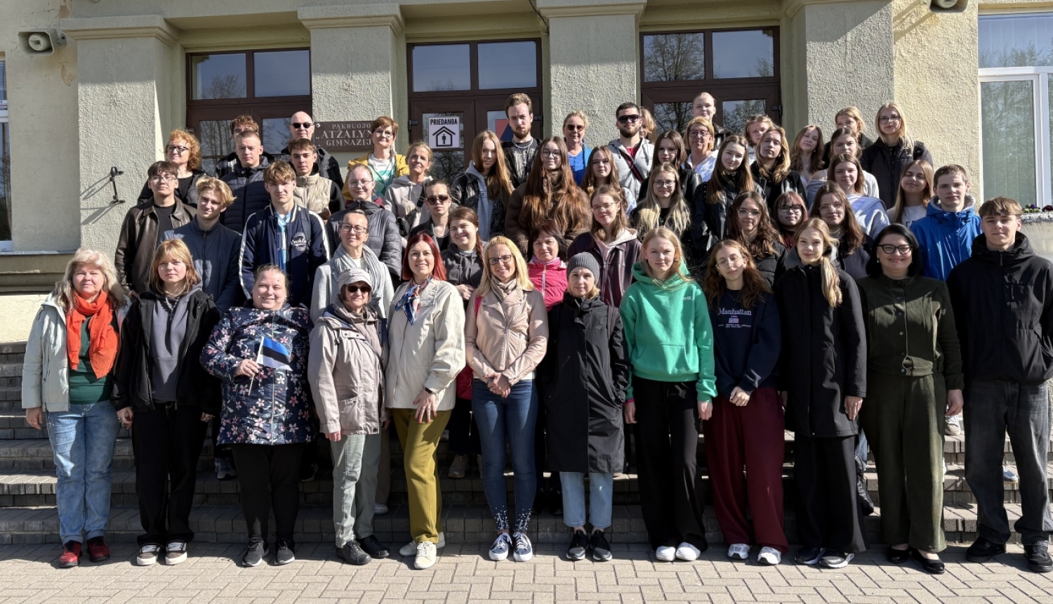 Attēlā redzama liela cilvēku grupa, kas pozē kopīgai fotogrāfijai pie ēkas ieejas. Grupā ir dažāda vecuma cilvēki – gan jaunieši, gan pieaugušie. Visi stāv uz kāpnēm un laukuma ēkas priekšā saulainā dienā. Daudzi ir ģērbušies jakās un mēteļos, kas liecina par vēsāku laiku. Fotogrāfija izskatās kā skolas, organizācijas vai kopienas pasākuma kopbilde.