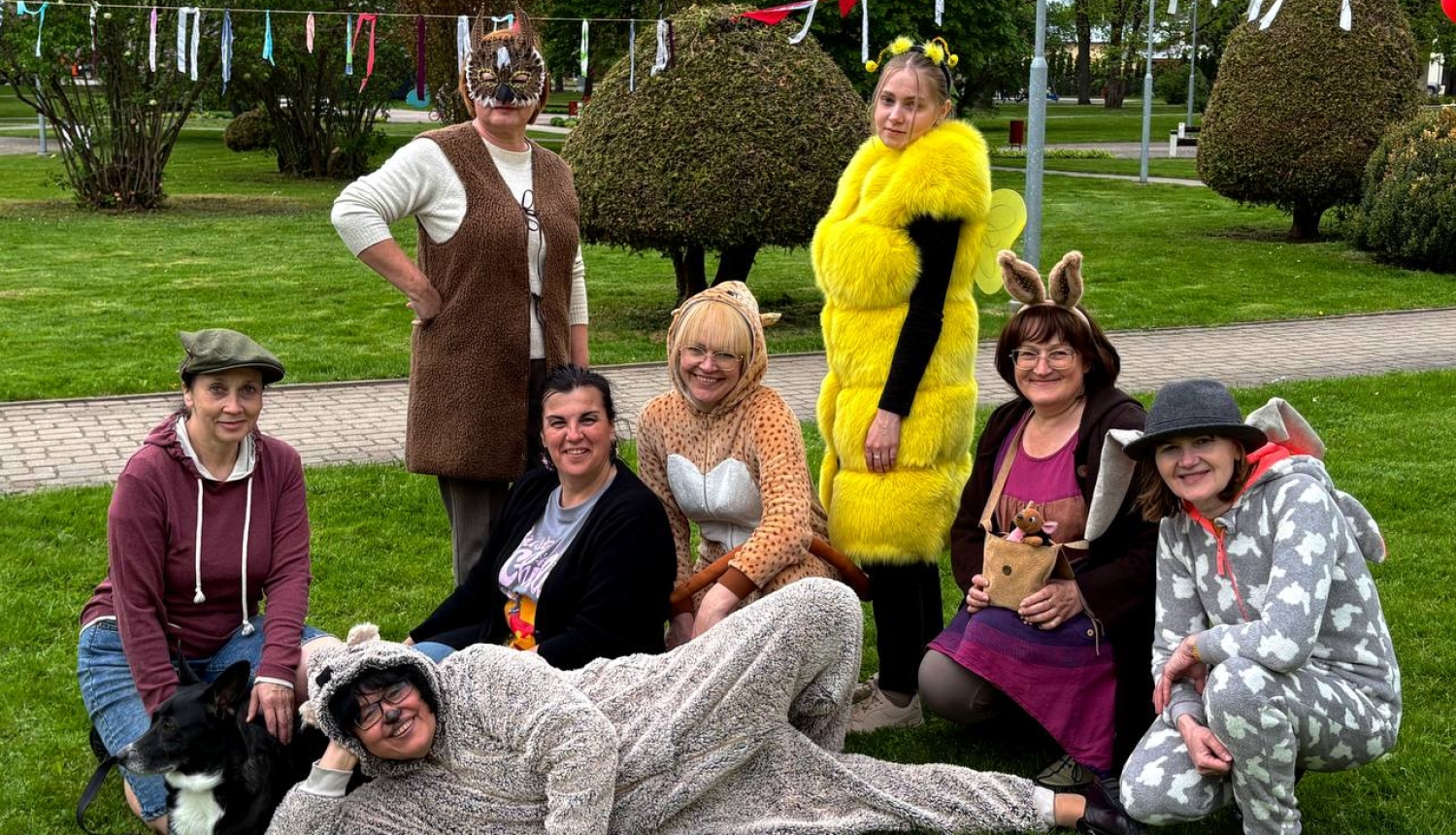 Grupa cilvēku parkā pozē kopīgai fotogrāfijai, tērpušies dažādos dzīvnieku un pasaku tēlu kostīmos. Daļa sēž un tup zālienā, citi stāv, radot rotaļīgu un priecīgu noskaņu. Fonā redzami dekorējumi un sakopts parks. Blakus grupai atrodas arī suns.