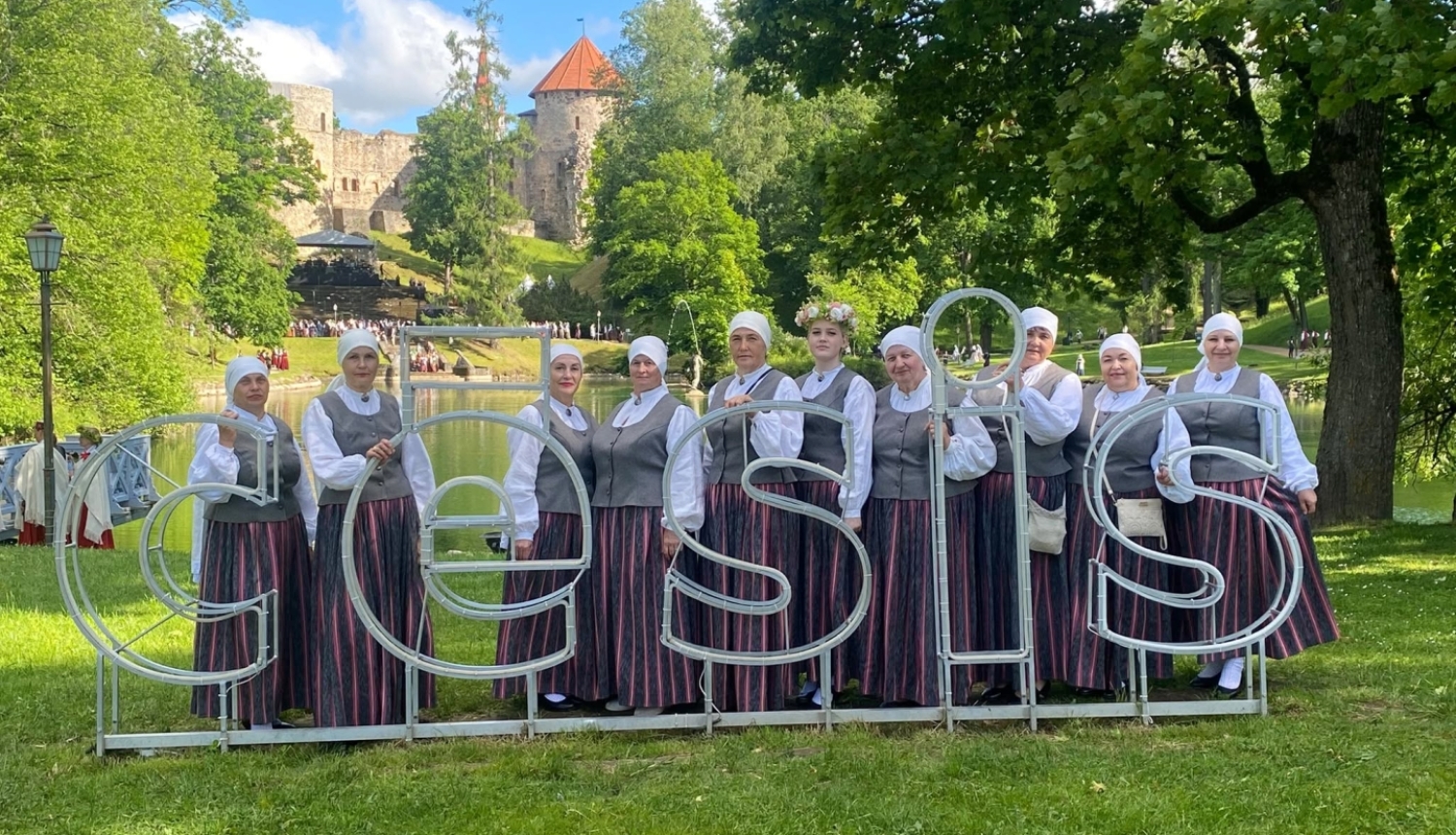 Sieviešu koris tautas tērpos pozē Cēsu Pils parka zālienā aiz liela, balta vides objekta — burtiem "CĒSIS". Fonā redzams dīķis un Cēsu viduslaiku pils drupas zaļumu ieskautā kalnā.