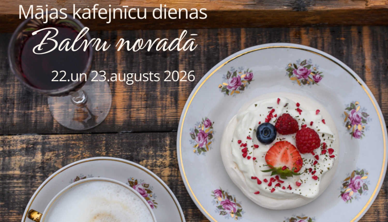 Attēlā redzama koka virsma un uz tā novietota pavlova kūciņa ar ogām, balta kafija un sarkanvīna glāze.